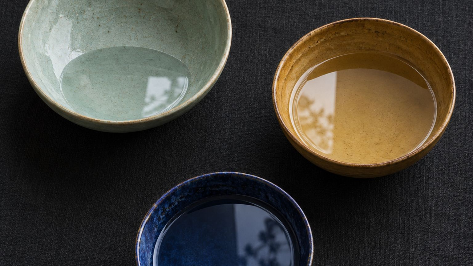 Three shallow ceramic bowls in different glazes - pale celadon, warm ochre, deep cobalt - stand on a matte linen surface, each holding water at a visibly different level, shot straight down from directly overhead. Soft diffused daylight makes each water surface a distinct ellipse of reflection, and the empty space above the lowest waterline is the clearest thing in the frame. No text, numbers, logos, people, hands, or faces.